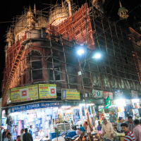A busy night market scene in India features a historic building under renovation, its ornate facade wrapped in scaffolding. Bright storefronts at street level display colorful signs like "Hajira Stores." People bustle around, and a few vehicles, including a bicycle and motorbikes, are parked nearby.