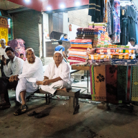 Three men sit on a wooden bench in a vibrant Indian market. Behind them, stacks of folded clothing and fabrics are displayed. Vendors and mannequins mingle amidst the colorful textiles. The setting is lively, blending traditional and modern attire against a rich cultural backdrop.