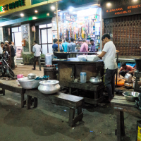 Street scene in India showing people in front of a restaurant. Two men wash dishes under dim lights while motorcycles are parked nearby. A shop with colorful items is visible in the background, and the street sign reads "ZAKARIA STREET." There’s a no-entry traffic sign on the left.