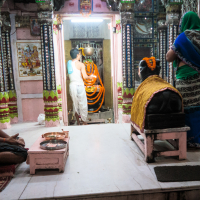 In a vibrant Indian temple adorned with garlands and colorful fabrics, a man worships before an idol while others observe. The interior features pink walls and intricate columns. Nearby, a woman in a green and blue sari stands, as a man sits cross-legged on the white marble floor.