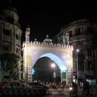 A night view of an illuminated gateway with intricate designs, reminiscent of India's architectural charm, resembling an arch with minarets. The lively scene includes people walking and cars passing by, while the dimly lit buildings around accentuate the brilliance of the bright arch.