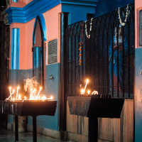 Candles are lit on stands in front of a building with blue and orange walls, reminiscent of vibrant India. The entrance features arched windows, and garlands hang on black metal bars. Plaques or signs adorn the wall, while the dim lighting creates a warm and peaceful ambiance.