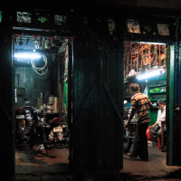 In a dimly lit garage reminiscent of India's bustling backstreets, two open doors reveal motorcycles inside. A man in a striped shirt stands nearby, while another sits inside. The walls are adorned with tools, and fluorescent lights illuminate the space, casting shadows over posters on the exterior wall.