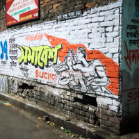 A brick wall on a dimly lit street in India features vibrant graffiti with Bengali text. The artwork shows two figures reaching towards a red flag. Surrounding slogans and text, partially obscured or faded, cover the white-painted section of the wall.