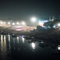 A nighttime riverside scene in India showcases illuminated ghats reflected in the water. Small boats are anchored along the riverbank, while trees and a few buildings, including a temple with a pointed tower, stand in the background. The area is bathed in soft, ambient light.