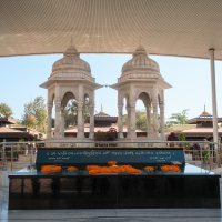 A serene memorial site in India features a black granite platform adorned with orange marigold flowers. Two intricately carved white stone pavilions stand behind it, under a modern, curved roof. Trees and additional structures surround the area, creating a peaceful ambiance.