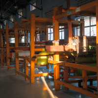 Wooden handlooms are arranged in a row in an indoor space, illuminated by sunlight streaming through large windows. Various levers, strings, and fabric components create intricate machinery used for weaving textiles, reminiscent of traditional looms found in India. The floor is smooth and shiny.