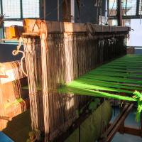 A traditional wooden loom from India set up with green threads ready for weaving. The loom is situated in a well-lit room, with sunlight filtering through large windows in the background. Beams of light highlight the intricate threads, showcasing the loom's detailed structure and craftsmanship.