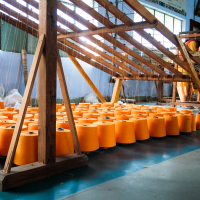 Orange spools of thread are neatly arranged in rows on a blue-painted factory floor in India. A wooden loom stands in the background, and a worker can be seen operating machinery. The well-lit setting highlights the orderly setup and equipment essential for textile production.