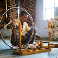 A person in a yellow and white checkered shirt and plaid pants operates a traditional spinning wheel on a blue floor indoors. The background, adorned with tapestries and a bright window, evokes the essence of India. The spinning wheel features large circular and smaller wooden parts, with text in Hindi partially visible in the background.