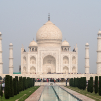 The image displays the iconic Taj Mahal of India, a white marble mausoleum with a large central dome, four minarets, and intricate detailing set against a hazy sky. In the foreground, a long rectangular reflecting pool is surrounded by landscaped gardens and pathways lined with cypress trees.
