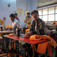 Four men are in a sewing workshop with two of them working at sewing machines on the front tables and two standing in the background. The workspace, reminiscent of traditional workshops in India, is filled with colorful fabrics, pictures on the wall, and a large window with blinds allowing sunlight to enter.