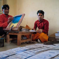 Two men sit on the floor in a room with a window, assembling printed booklets. One operates a printing press on a small table while the other organizes the booklets. Both wear traditional Indian attire and are deeply focused on their tasks, surrounded by piles of booklets and scattered papers.