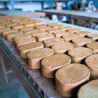 Rows of cylindrical clay pieces are neatly arranged on a wooden table in an Indian workshop setting. The smooth, brown surfaces reflect light from a window, creating a slight sheen. Shelves and various tools are blurred in the background, indicating a vibrant workspace for pottery or sculpture.