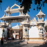The image shows an intricately designed temple facade in India, featuring white and gold architectural elements. The entrance is adorned with statues and detailed carvings. The clear blue sky and greenery frame the scene, highlighting the temple's grandeur and serene surroundings. Two people are walking towards the entrance.