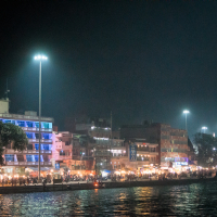 A night-time scene of a bustling riverside market in India, illuminated by streetlights and shop lights. The market consists of several buildings and stalls along the water, with a mix of modern and traditional architecture. Shoppers and vendors populate the lively street, reflecting in the river.