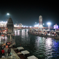 Night view of Har Ki Pauri ghat in Haridwar, India. The scene shows the Ganges River lined with illuminated temples and buildings. Devotees and visitors are gathered along the steps, engaging in rituals and activities. The river is flanked by a large clock tower in the background, epitomizing India's rich heritage.