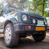 A green off-road vehicle parked on a dirt pathway surrounded by trees, likely somewhere in India. The front of the car features two large headlights and a license plate reading "UK07 TB 1735." A sign on the grille reads "WILD LIFE NATURE SAFARI," indicating it may be used for nature tours or safaris.