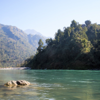 A serene river flows with clear turquoise water, bordered by rocky banks. Tall, lush green trees cover the hillside on the right, while layers of hazy mountains rise in the background under a clear blue sky. A bridge is faintly visible in front of the trees, spanning the river at a distance in India.