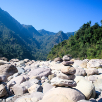 A serene landscape in India featuring a pile of balanced stones in the foreground, set against rocky terrain leading towards lush, green mountains. The sky above is clear and bright blue, enhancing the tranquil atmosphere of this mountainous valley.