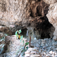 A rustic stone entrance to a cave with a stack of rocks at the forefront evokes the ancient charm of India. Hanging plants drape near the entrance, and the dimly lit interior adds to its mystique. The jagged, uneven walls and ceiling reveal slight hints of greenery peeking from the edges.