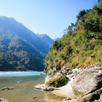 A serene riverside landscape in India with clear blue skies. The river flows gently, bordered by rocky and sandy shores. Lush green hills rise in the background under bright sunlight. The scene is tranquil, with clear water reflecting the greenery and sky.