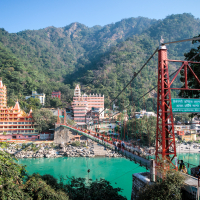A vibrant suspension bridge spans a green river, bustling with people crossing. On the left, intricately designed multi-story temples and colorful buildings typical of India line the riverbank. Lush green mountains rise in the background under a clear blue sky.