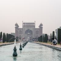 A wide-angle shot of a serene garden in India with a long rectangular reflecting pool leading to a grand Mughal-style gateway in the distance. The pool is adorned with ornate fountains, lush green cypress trees flank the pool, and people are visible near the gateway and poolside, some tending to the garden.