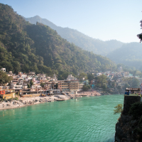 A calm turquoise river winds through a scenic valley surrounded by lush green mountains in India. On the left bank, a quaint town with colorful buildings sits nestled against the mountainside, while dense vegetation covers the slopes. Clear blue sky forms the backdrop of this serene landscape.