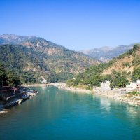 A serene river flows between two lush green mountainous terrains under a clear blue sky. On the right bank, small buildings and structures are visible nestled among the trees. On the river, a few people are engaging in water activities like rafting. This picturesque landscape could easily belong to India.