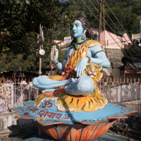 A vibrant statue of Lord Shiva in a meditative pose, painted blue and adorned with a tiger skin garment, is seated on a lotus pedestal. The deity holds a trident and has a serene expression. This iconic symbol of India is encircled by metal fencing with trees, buildings, and power lines in the backdrop.