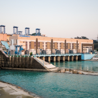 A hydroelectric power plant near a river in India. The facility features a large, rectangular building with multiple small towers on its roof. Concrete structures are used to channel water, and the water in the foreground appears calm, with a blue-green hue. Trees and power lines are visible in the background.