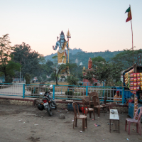 A large statue of Lord Shiva stands in the background with hills and trees behind it, evoking a serene scene typical of India. In the foreground, a few plastic chairs and tables are near a small, blue shop selling various packaged goods. A motorcycle is parked nearby, and a flag flutters on a pole to the right.