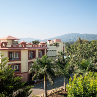 A serene view of a multi-story building with red and cream-colored walls, typical of India, is surrounded by lush green palm trees and dense foliage. A clear road runs beside the building. In the background, rolling hills frame the scene under a clear blue sky.