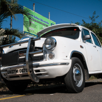 A white vintage car with a prominent front grille and bumper guard is parked on a sunny road in India. The car has a license plate marked "UA07 M 0674." In the background, there's lush greenery and a large signboard under the bright blue sky.
