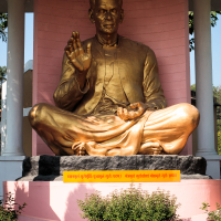 A large golden statue depicting a seated figure in a meditative pose with one hand raised and the other resting on the knee is positioned under a canopy. Set against a pink backdrop, with India’s rich culture reflected in a plaque featuring text in Hindi at the bottom, it's surrounded by plants and flowers.