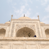 A low-angle view of the Taj Mahal's main dome and ornate marble facade against a partly cloudy sky in India. The central arch showcases intricate floral designs and inlaid patterns. Two people are visible on the terrace, one in camouflage attire and the other wearing a blue outfit.