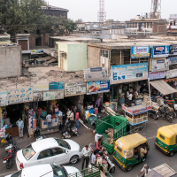 A bustling street scene in an urban area of India with a line of shops selling various goods. Auto rickshaws and a white car are seen on the road, along with numerous pedestrians. Overhead cables stretch across the scene, and buildings behind the shops are visible. The area is lively with activity.