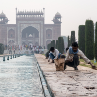 Three men clean and sweep the edge of a long, narrow water feature lined with fountains in front of a grand, large red and white gateway with arches and domes. Tall cypress trees flank the water feature, while tourists explore in the background. The hazy sky adds an ethereal touch to this iconic Indian scene.