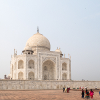 The image showcases the Taj Mahal, a white marble mausoleum located in Agra, India. It features a large central dome flanked by four minarets at each corner. Tourists are gathered in the foreground on a broad stone plaza, taking photos and admiring the historic structure on a clear day.