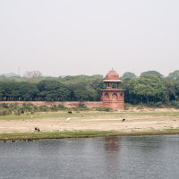 A solitary red sandstone pavilion stands by a river, surrounded by lush green trees, reminiscent of pastoral India. In the foreground, the river has a grassy bank with cattle grazing. The overcast sky blends softly into the distant vegetation, creating a calm and serene scene.
