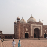 People visit a historic, intricately-designed mosque in India, with multiple beige domes and minarets in the distance. The red and white structure features detailed carvings and large archways. The foreground reveals a paved courtyard with visitors in traditional attire, capturing the architectural grandeur.