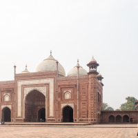 The image shows the Taj Mahal mosque in India, made of red sandstone with intricate white marble inlay. It features three domes and four minarets. Three people walk in the foreground on the expansive courtyard, two wear beige and red and one in red and blue. The sky is clear and light.