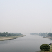 A serene river flows calmly under a hazy sky, bordered by green trees and vegetation. The foggy atmosphere softens the distant view, reminiscent of picturesque riverbanks in India. A pagoda and small buildings are visible on the right bank, exuding tranquility and natural beauty with minimal human activity.