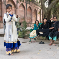 A young girl in traditional dress stands in the foreground, fingers near her mouth, looking contemplative. Behind her, five women, also in traditional attire, are seated on a bench under a tree. The setting appears to be an outdoor area in India with stone pathways and historic reddish-brown arches.
