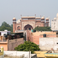View of the Taj Mahal from a distance, with a foreground of varied rooftops and buildings. The image prominently features the intricately detailed gateway leading towards India's iconic Taj Mahal, with its white marble dome visible in the background against a hazy sky.