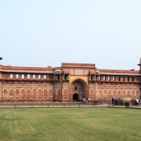 The image shows a panoramic view of Agra Fort, a UNESCO World Heritage site in India. The red sandstone building features intricate white marble inlay work, arched windows, and two large domes. In front of the fort is a well-maintained lawn with a few visitors walking along a pathway.