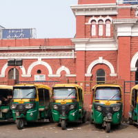 Six colorful auto rickshaws are parked in a row in front of a historic red brick building with ornate white trim, typical of India's rich architecture. The building has arched windows and a blue sign with text in an Asian script. A clutter of items and some debris are visible around the rickshaws.