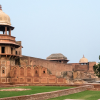 The image showcases Agra Fort in India. Visible are historic red sandstone structures, arched entrances, and an ornate dome. The fort's walls are interspersed with detailed carvings and windows. Lush green lawns and a large tree add contrast to the ancient architecture under India's clear sky.