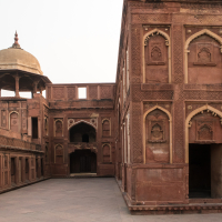 A large courtyard enclosed by reddish-brown stone buildings adorned with detailed carvings and arched windows. The building on the left features a prominent domed pavilion atop a multi-arched structure, reminiscent of India's rich architectural heritage with its intricate patterns and historic design elements.